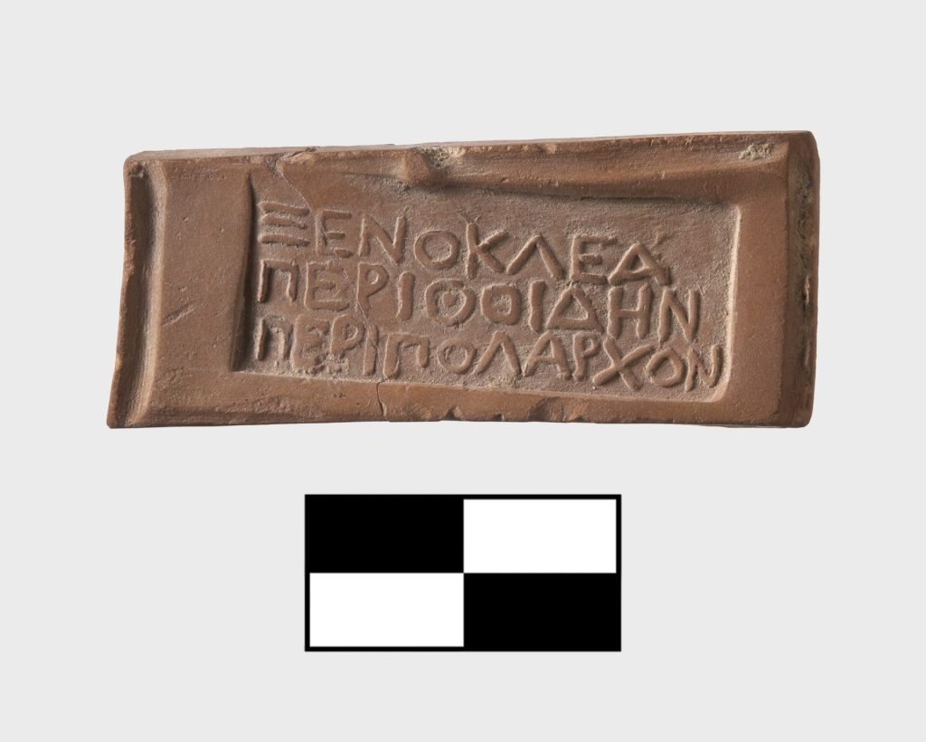 Rectangular terracotta plaque bearing a Greek inscription incised in multiple lines.Black-and-white scale graphic used to indicate measurement for archaeological documentation.